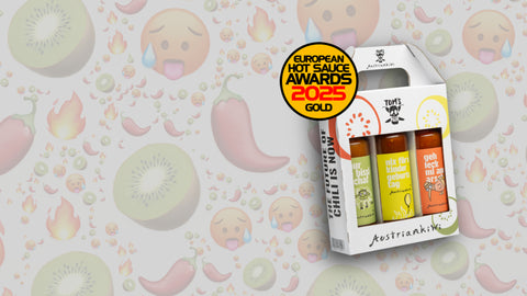 3er-Set scharfer Chilisaucen von AustrianKiwi und TOM'S HOT STUFF in bunter Verpackung, vor Hintergrund mit Kiwi-Scheiben, Chilischoten, Flammen und schwitzenden Emoji-Gesichtern.